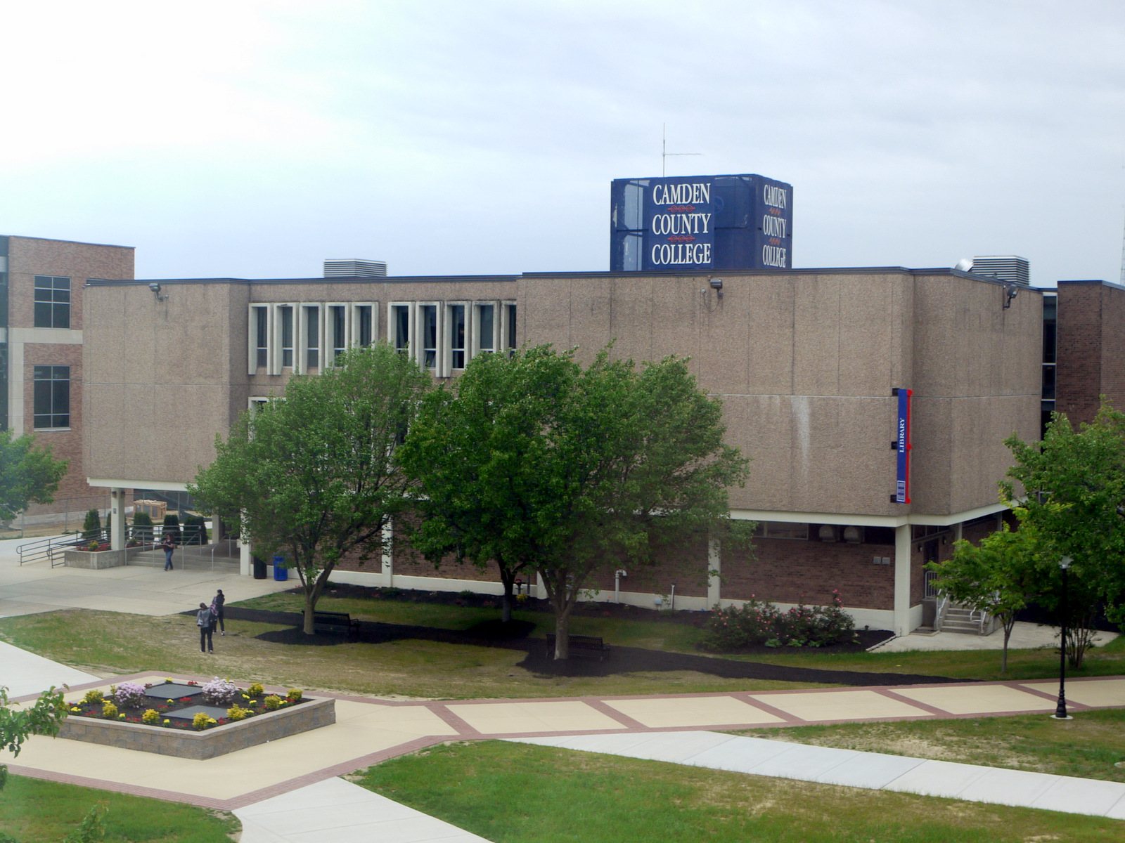 A tan concrete library building with a blue rooftop sign reading Camden County College, with trees and brick walkways in front