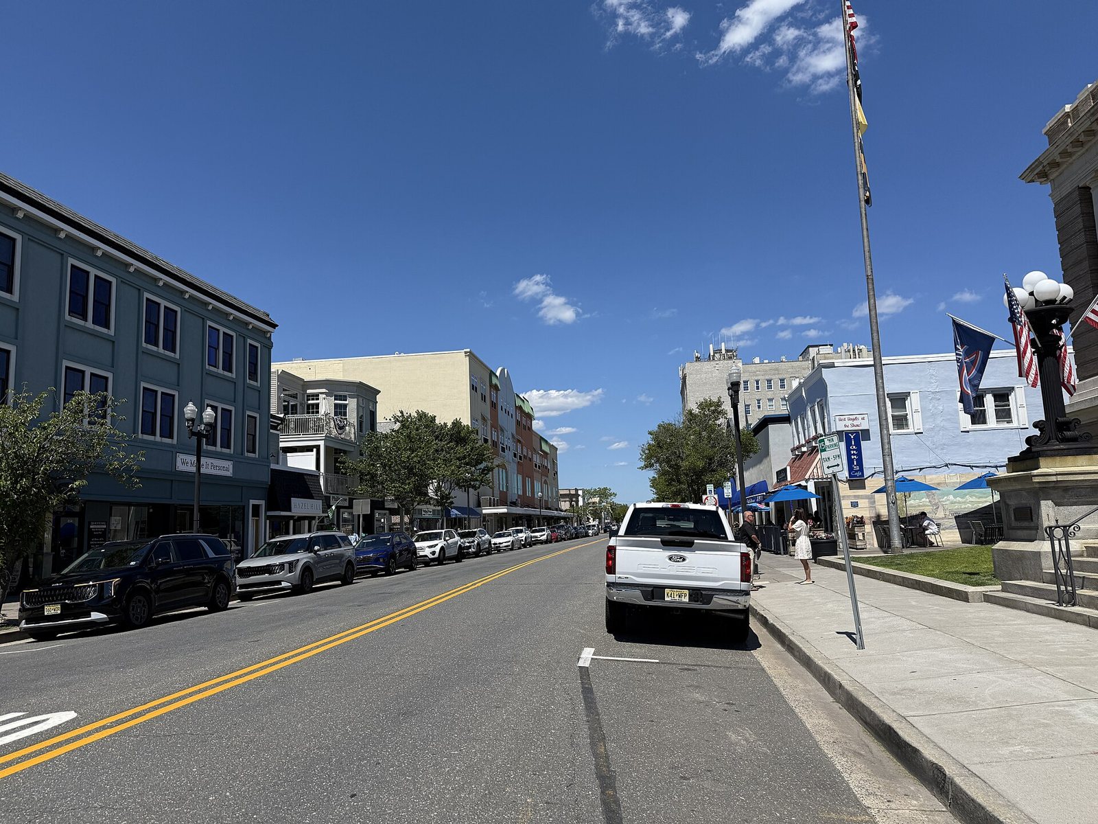 A sunny downtown street lined with shops and parked cars, with flags and a lamppost beside a stone building on the right
