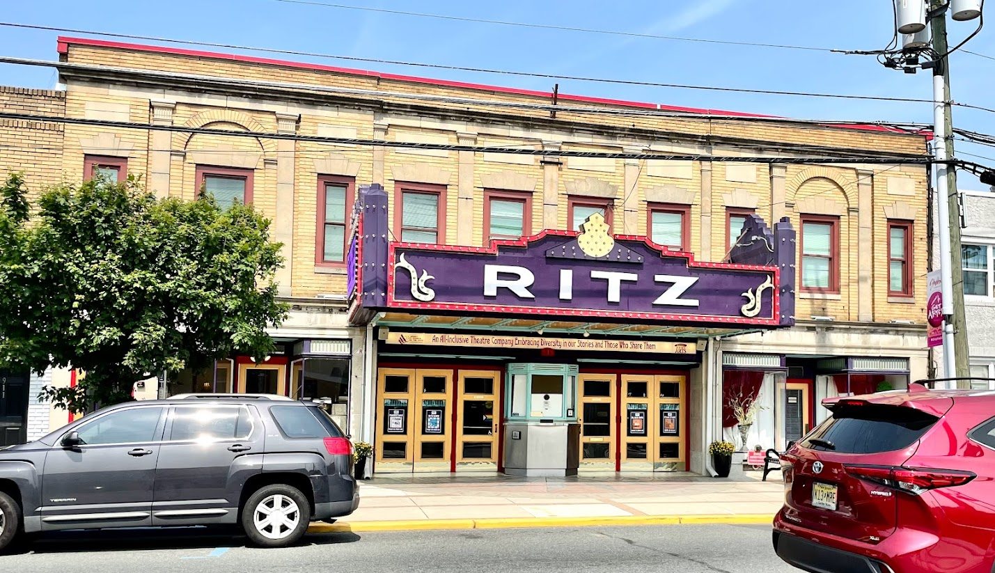 A tan brick theater building with a purple marquee reading RITZ above a row of gold-framed doors and a ticket booth