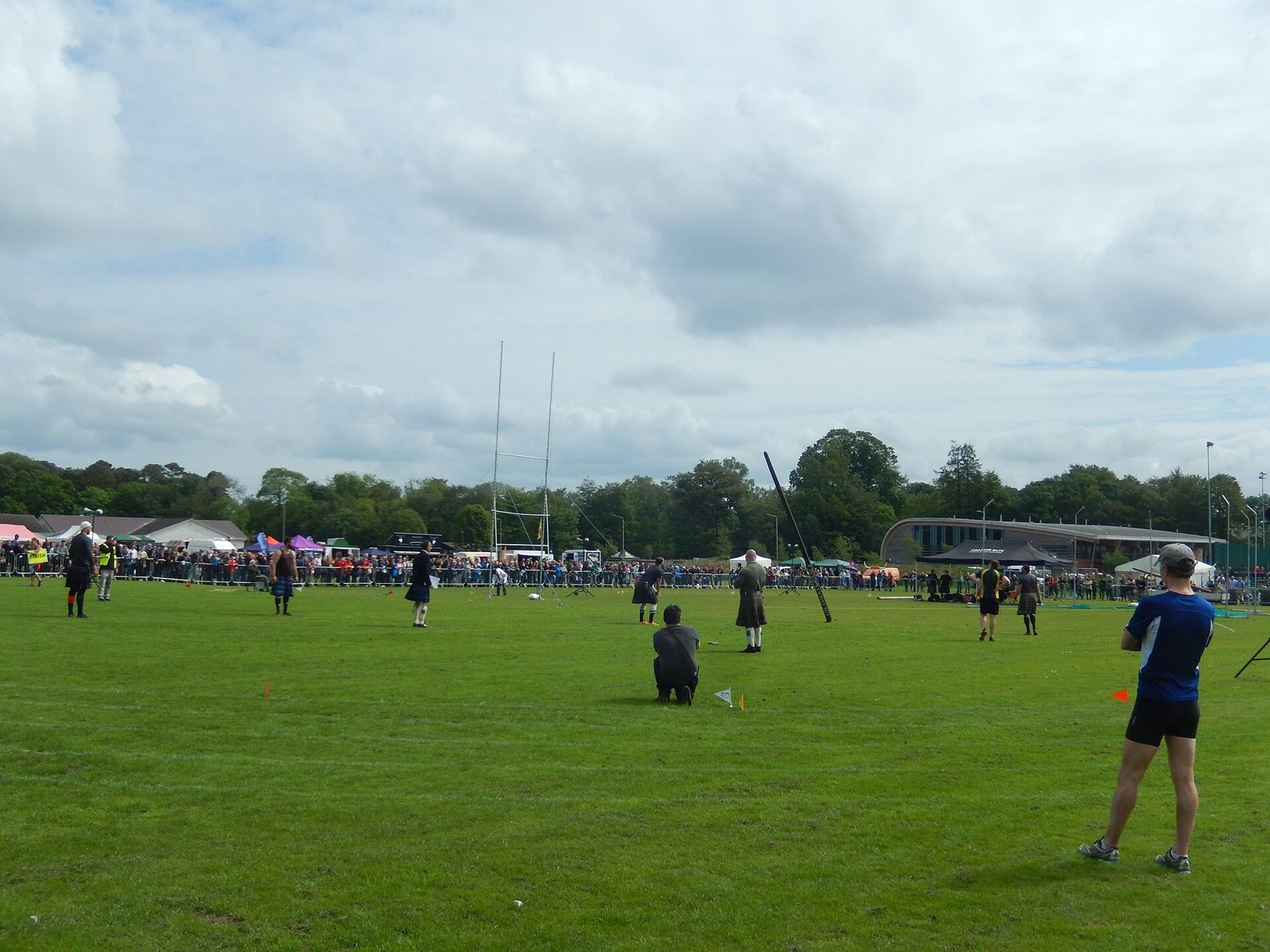 A caber in mid-air above a grass field as kilted athletes and a fenced-off crowd look on