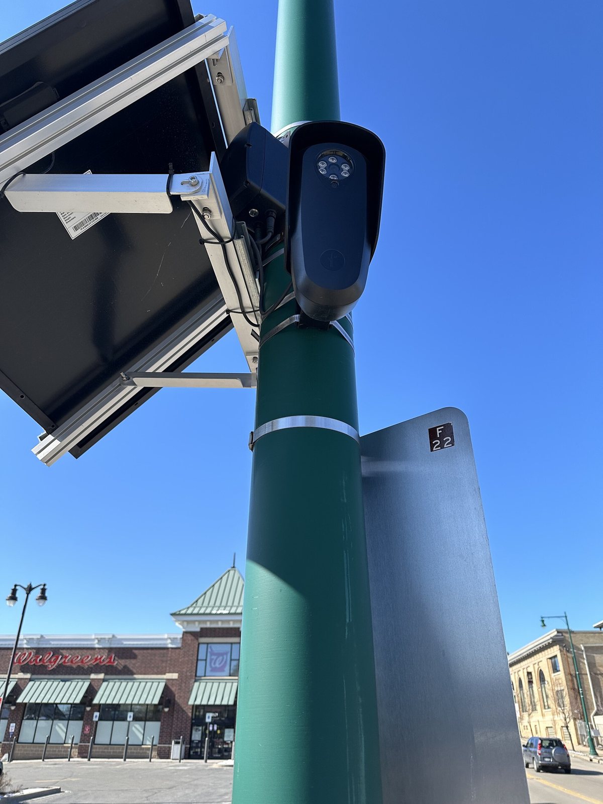 A black Flock Safety license-plate camera and solar panel mounted on a green street pole under a blue sky